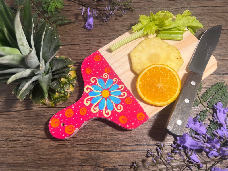 Pu&ograve; includere: Un tagliere di legno dipinto di rosa e blu con un motivo floreale. Il tagliere ha un manico ed &egrave; decorato con punti arancioni e bianchi. Un ananas, un'arancia e fette di ananas sono sul tagliere. Un coltello &egrave; sul tagliere.