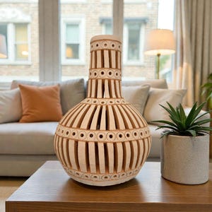 Op de afbeelding: Een decoratieve, gebroken witte keramische vaas met een uniek ontwerp van verticale uitsparingen en cirkelvormige accenten. De vaas staat op een houten salontafel, met een kleine plant in een pot rechts. De vaas is ongeveer 46 cm hoog.