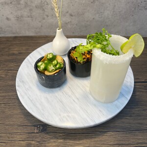 May include: A white marble serving tray with a white vase, a glass of white liquid with a salted rim, a small bowl of green peppers and chips, and a small bowl of brown food topped with green herbs.