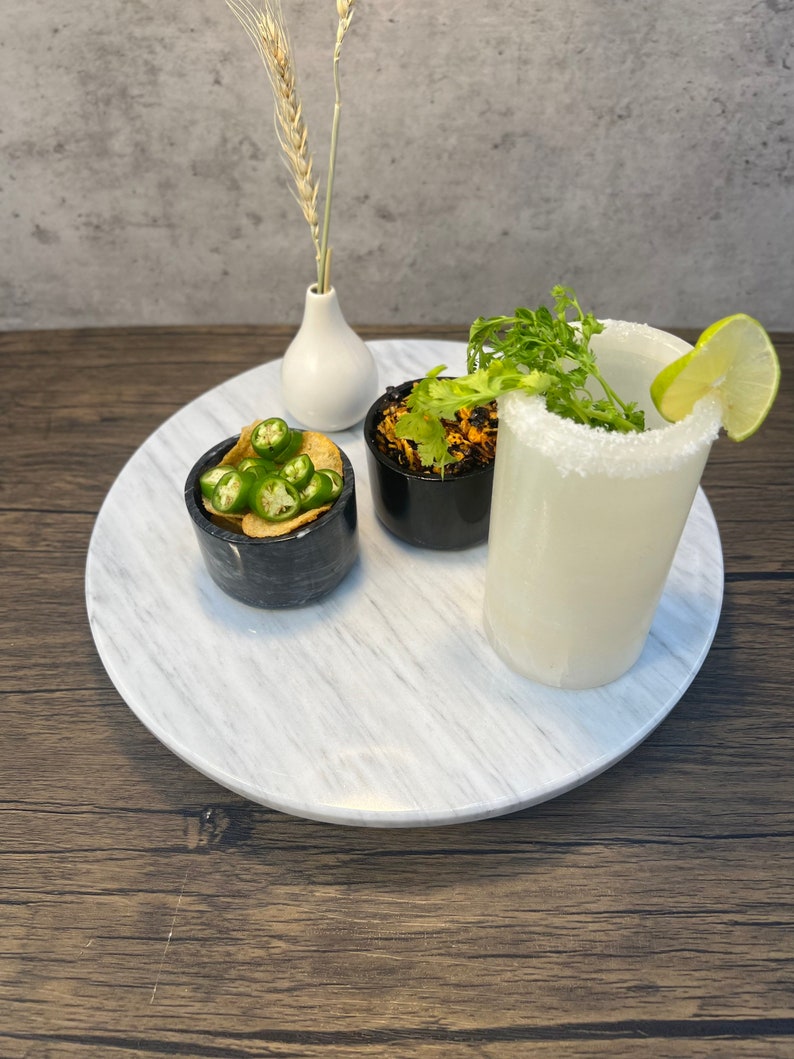 May include: A white marble serving tray with a white vase, a glass of white liquid with a salted rim, a small bowl of green peppers and chips, and a small bowl of brown food topped with green herbs.
