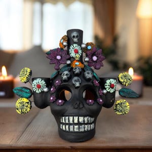 May include: Black ceramic skull candle holder with three candle cups and floral decorations. The skull has white teeth and is decorated with small skulls and flowers in various colors.