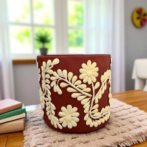 Könnte beinhalten: Ein terrakottafarbener Keramik-Blumentopf mit cremefarbenem Blumendesign. Das erhabene Reliefmuster zeigt Gänseblümchen und Blattranken. Der Topf steht auf einer neutralfarbenen Matte.