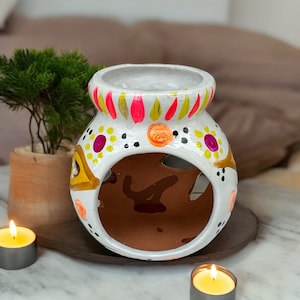 May include: A white ceramic essential oil diffuser with colorful hand-painted accents. The diffuser features a large opening and is decorated with pink, orange, and yellow designs. Two lit candles are in the foreground.