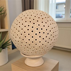 Op de afbeelding: Een beige bolvormige decoratieve lamp met een patroon van gelijkmatig verdeelde cirkelvormige uitsparingen. De lamp staat op een witte voet en wordt tentoongesteld op een lichtgekleurd houten blok. Groene planten in witte potten staan op de achtergrond.