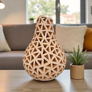 Op de afbeelding: Een decoratieve, lichtgekleurde vaas met een geometrisch driehoekig patroon. De vaas staat op een houten tafel, naast een kleine plant in een pot. De achtergrond omvat een grijze bank en kussens.
