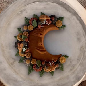 Puede incluir: Una luna creciente marrón con una cara esculpida en ella, rodeada de una corona de flores y mariposas coloridas. La luna está sobre una superficie de mármol blanco.