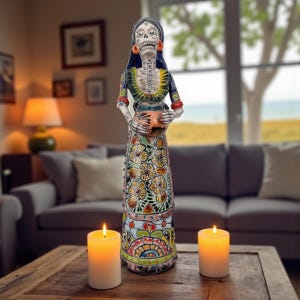 May include: A tall, colorful Day of the Dead figurine. The ceramic sculpture depicts a female skeleton figure holding a pot. The figure is adorned with intricate floral and geometric patterns in various colors. Orange earrings and a black head covering complete the look.