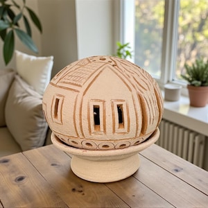 Op de afbeelding: Een beige keramische decoratieve lamp in de vorm van een bol met uitgesneden huisontwerpen en rechthoekige ramen. De lamp staat op een bijpassende voet. De lamp staat op een houten tafel.