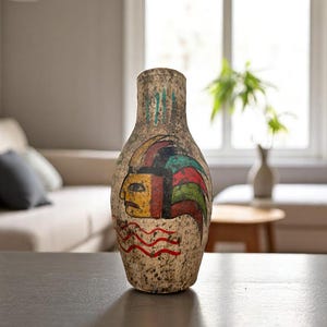 May include: A tall, textured vase with a painted design featuring a face with a colorful headdress. The vase has a beige and brown color scheme with red, yellow, green, and blue accents. The vase is displayed on a dark surface.