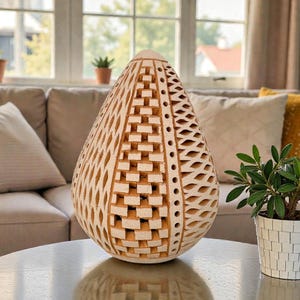 Op de afbeelding: Een decoratieve, druppelvormige houten sculptuur met ingewikkelde uitgesneden patronen. De lichtbruine sculptuur staat op een ronde tafel. Een kleine plant in een pot staat ernaast. De achtergrond toont een bank en een raam.