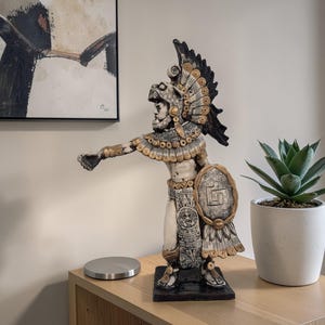 May include: A detailed statue of an Aztec warrior, featuring intricate details in black, gold, and cream colors. The warrior is adorned with a feathered headdress, shield, and ornate clothing. The statue is displayed on a wooden surface, next to a potted succulent.
