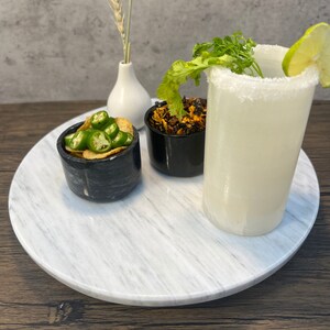 May include: A white marble serving tray with three small black bowls. One bowl contains green jalapeno peppers, another contains dried red chili flakes, and the third bowl is empty. A tall glass of white liquid with a salted rim and a lime wedge is on the tray.