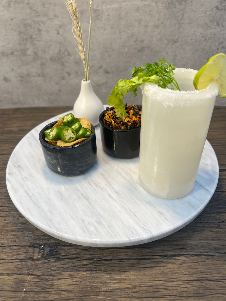 May include: A white marble serving tray with three small black bowls. One bowl contains green jalapeno peppers, another contains dried red chili flakes, and the third bowl is empty. A tall glass of white liquid with a salted rim and a lime wedge is on the tray.