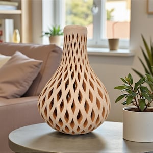 Op de afbeelding: Een beige decoratieve vaas met een uniek opengewerkt ontwerp. De vaas heeft een bolvormig lichaam en een smalle hals, met een reeks ruitvormige uitsparingen. Hij staat op een grijze tafel naast een kleine potplant.