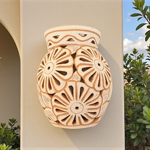 Op de afbeelding: Een beige keramische vaas met een bloemenontwerp. De vaas heeft een afgeronde vorm met uitgesneden bloempatronen en een decoratieve bovenkant. De vaas is bevestigd aan een lichtgekleurde muur, met groen in de achtergrond.