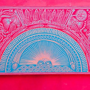 May include: A framed artwork with a colorful, intricate design. The artwork features a blue and white sun with rays emanating from it, surrounded by a red background with white and silver stylized figures and patterns.