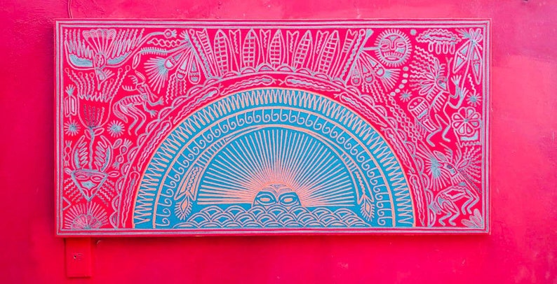 May include: A framed artwork with a colorful, intricate design. The artwork features a blue and white sun with rays emanating from it, surrounded by a red background with white and silver stylized figures and patterns.
