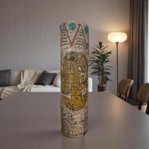 May include: A tall, cylindrical vase with a textured, speckled surface. The vase features a detailed, colorful design with yellow, blue, and brown tones. The artwork appears to depict a stylized figure or face, with geometric patterns above.