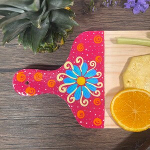 Pu&ograve; includere: Un tagliere in legno dipinto di rosa con un motivo floreale blu e giallo. Il tagliere ha una piccola maniglia ed &egrave; decorato con punti bianchi. Il tagliere &egrave; appoggiato su una superficie di legno con un ananas, sedano, arancia e un pezzo di ananas sopra.