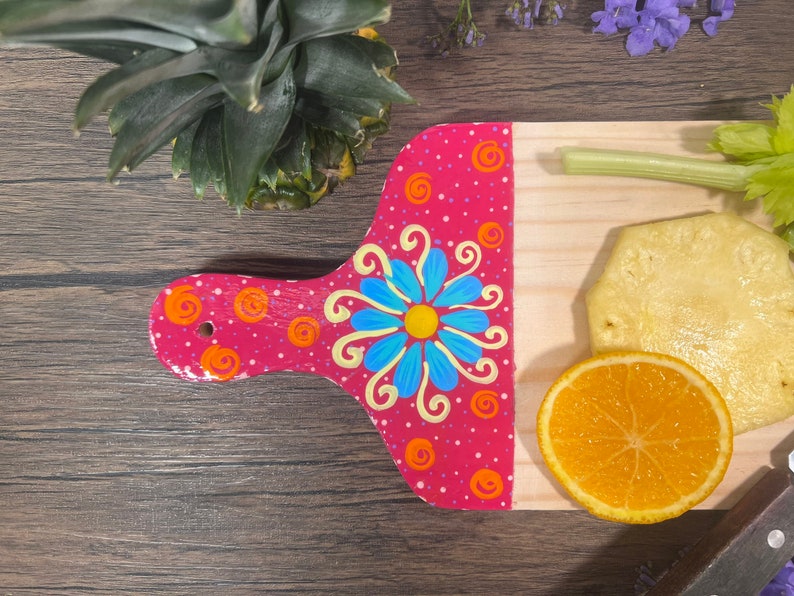 Pu&ograve; includere: Un tagliere in legno dipinto di rosa con un motivo floreale blu e giallo. Il tagliere ha una piccola maniglia ed &egrave; decorato con punti bianchi. Il tagliere &egrave; appoggiato su una superficie di legno con un ananas, sedano, arancia e un pezzo di ananas sopra.