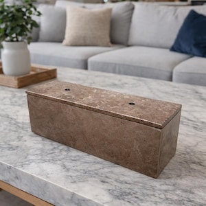May include: A rectangular brown marble box with a lid, featuring two small circular holes on top. The box is placed on a marble coffee table with a wooden frame, in a living room setting.