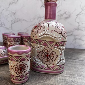 Puede incluir: Un juego de licor de cer&aacute;mica con una botella con tap&oacute;n y varios vasos de chupito. El juego est&aacute; pintado con un dise&ntilde;o floral en tonos rosa, rojo y blanco sobre un fondo beige. La botella y los vasos tienen un patr&oacute;n decorativo.