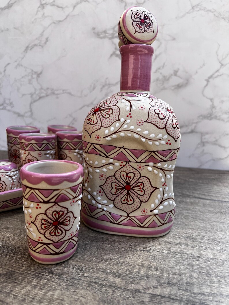 Puede incluir: Un juego de licor de cer&aacute;mica con una botella con tap&oacute;n y varios vasos de chupito. El juego est&aacute; pintado con un dise&ntilde;o floral en tonos rosa, rojo y blanco sobre un fondo beige. La botella y los vasos tienen un patr&oacute;n decorativo.