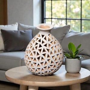 Op de afbeelding: Een witte decoratieve vaas met een geometrisch driehoekpatroon. De vaas staat op een ronde houten tafel naast een kleine potplant. De vaas heeft een brede opening en een textuur.