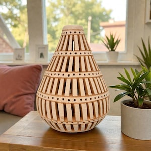 Op de afbeelding: Een beige, decoratieve lantaarn met een uniek opengewerkt ontwerp. De lantaarn heeft een afgeronde basis en een taps toelopende bovenkant, met verticale uitsparingen en gestippelde accenten. Hij staat op een houten tafel, naast een kleine potplant.