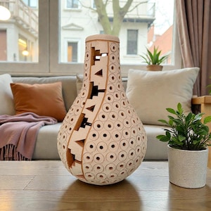 Op de afbeelding: Een decoratieve, lichtgekleurde houten vaas met een uniek ontwerp. De vaas heeft een bolvorm met een reeks kleine, gelijkmatig verdeelde cirkelvormige uitsparingen. Het ontwerp omvat geometrische vormen en uitgesneden patronen. De vaas staat op een houten tafel.