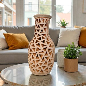Op de afbeelding: Een beige keramische vaas met een geometrisch driehoekpatroon. De vaas staat op een marmeren tafel, met een grijze bank en decoratieve kussens op de achtergrond. Een kleine plant in een pot staat in de buurt.