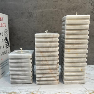 May include: Three white marble pillar candles of varying heights. The candles have a textured, ribbed design and are arranged in a row on a white marble surface.