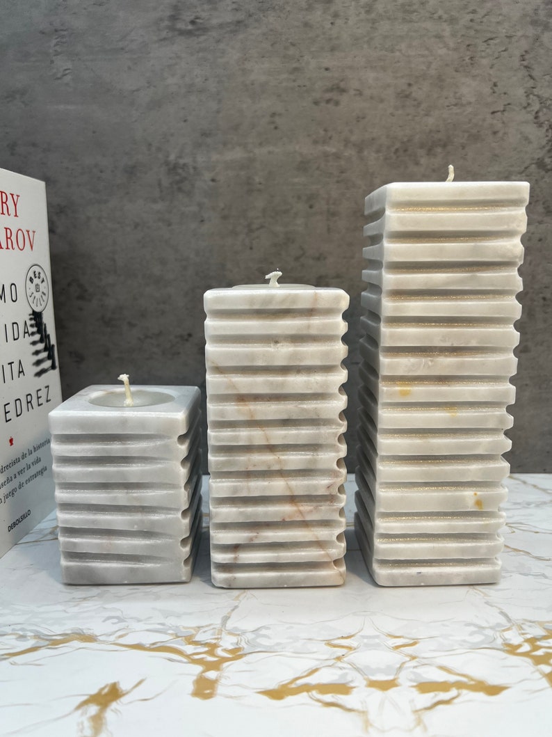 May include: Three white marble pillar candles of varying heights. The candles have a textured, ribbed design and are arranged in a row on a white marble surface.