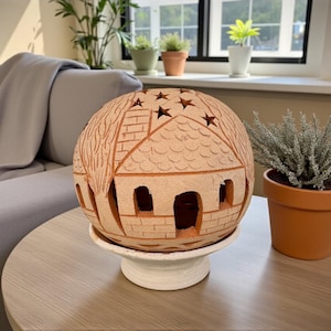 Op de afbeelding: Een bolvormige, terracotta-kleurige decoratieve lantaarn met een uitgesneden huisontwerp, inclusief ramen en een dak, staat op een witte sokkel. De lantaarn heeft stervormige uitsparingen. Een potplant staat naast de lantaarn.