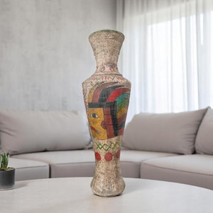May include: Tall, textured vase with a painted face design. The vase features a beige background with a colorful face design in yellow, red, and green. The vase is a decorative home accent.