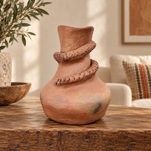 May include: A terracotta-coloured clay vase with a wide base and a narrow neck. A textured, snake-like coil wraps around the neck and body of the vessel. The surface features natural variations in colour, including patches of dark grey and reddish-brown tones.