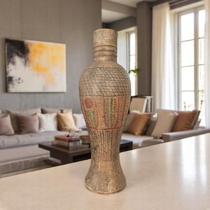 May include: A tall, textured vase with a narrow neck and a wide base. The vase features a geometric pattern and bands of red, green, and brown colors. The vase is displayed on a light-colored countertop in a living room setting.