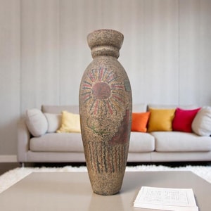 May include: A tall, textured vase with a wide base and a narrower neck. The vase is adorned with a sun-like design in red and brown, with additional colorful patterns. It stands on a light-colored table in front of a sofa.