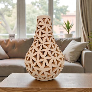 Op de afbeelding: Een beige, decoratieve vaas met een uniek bloemenpatroon. De peervormige vaas staat op een houten tafel. De achtergrond omvat een bank, planten en een raam, wat een warme sfeer creëert.