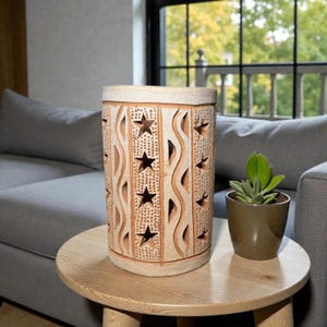 Op de afbeelding: Een cilindrische, beige keramische lantaarn met steruitsparingen en golvende patronen. De lantaarn staat op een ronde houten tafel naast een kleine vetplant in een pot. De achtergrond omvat een grijze bank en een raam.