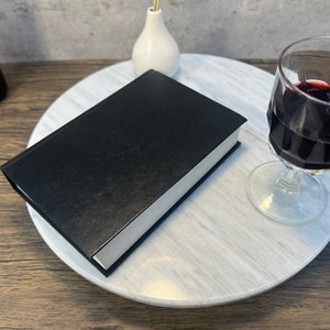 May include: A black leather-bound book on a white marble turntable with a glass of red wine and a white vase.