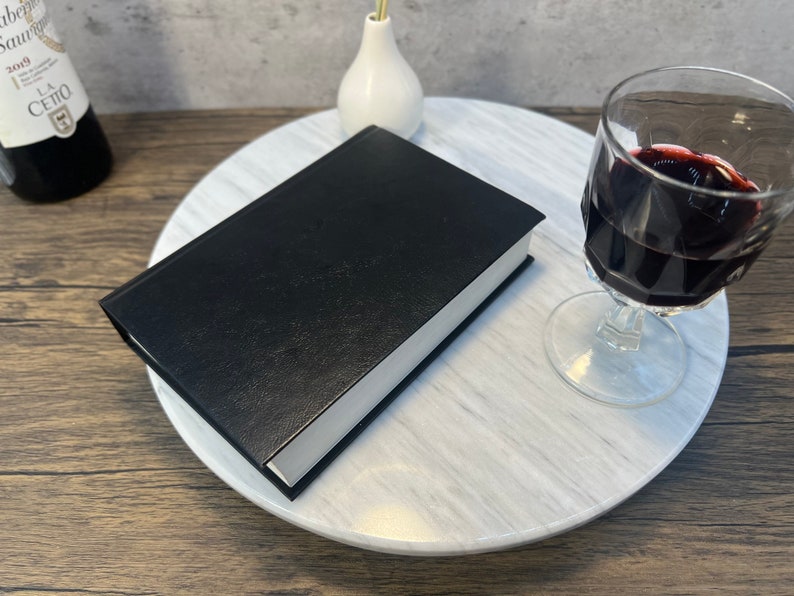 May include: A black leather-bound book on a white marble turntable with a glass of red wine and a white vase.