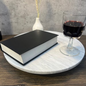 May include: A white marble cake stand with a black book and a glass of red wine on top. The stand is on a wooden surface.