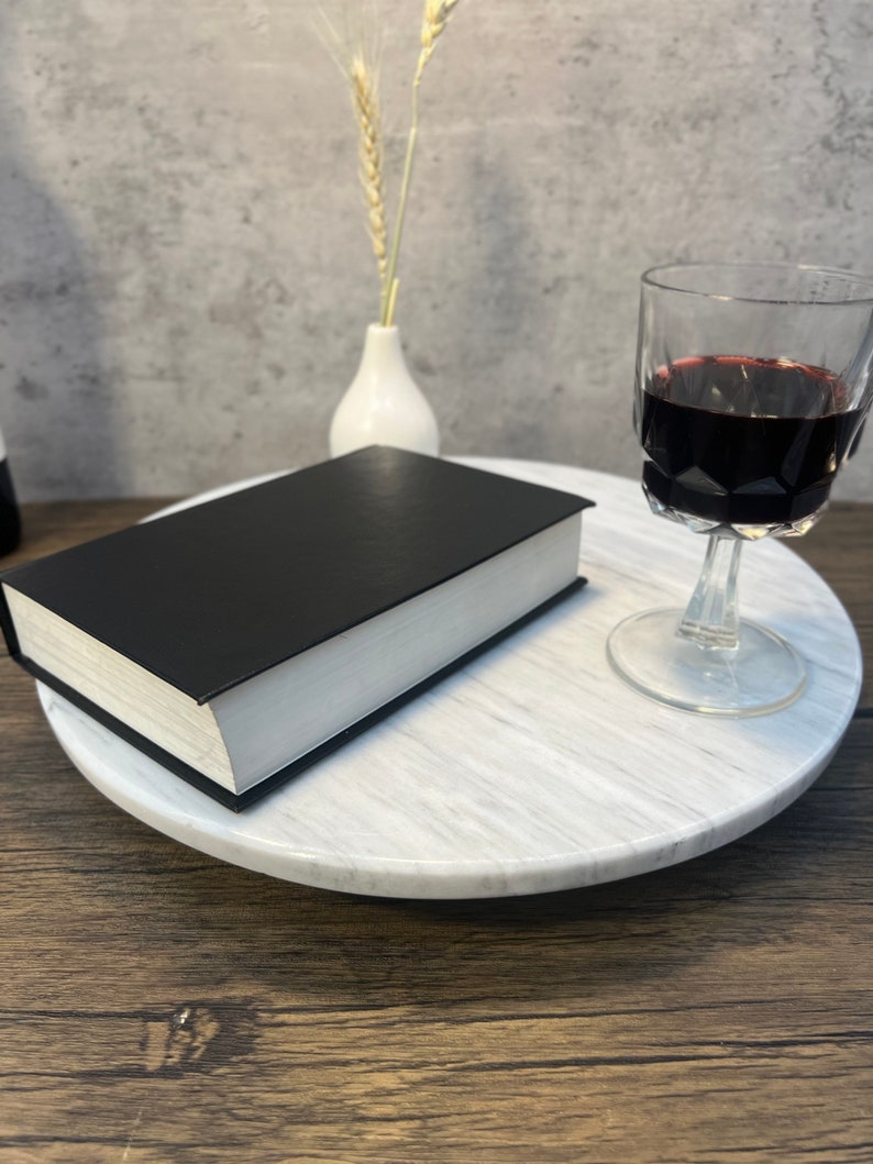 May include: A white marble cake stand with a black book and a glass of red wine on top. The stand is on a wooden surface.