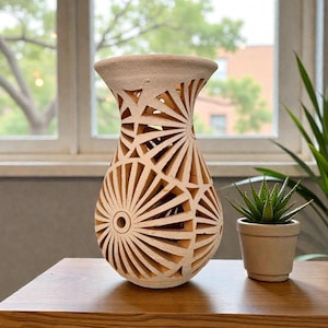 Könnte beinhalten: Eine beige, geschnitzte Keramikvase mit breitem Boden und ausgestelltem Oberteil. Die Vase weist ein geometrisches Sonnenstrahlenmuster auf, das durch die Keramik geschnitten ist. Rechts von der Vase steht eine kleine Topf-Sukkulente.