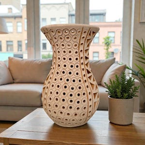 Op de afbeelding: Een beige decoratieve vaas met een uniek ontwerp van cirkels en driehoeken die in het oppervlak zijn uitgesneden. De vaas staat op een houten salontafel, met rechts een kleine plant in een pot. Een bank en een raam zijn op de achtergrond.