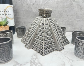 Puede incluir: Un decantador gris de cerámica con forma de pirámide maya con un juego de vasos de chupito a juego. El decantador tiene un diseño detallado de escaleras y elementos arquitectónicos. Una botella de Cabernet Sauvignon es visible en el fondo.