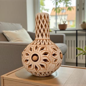 Könnte beinhalten: Eine beige, handgeschnitzte Keramikvase mit aufwendigen floralen und geometrischen Mustern. Die Vase hat einen bauchigen Körper und einen durchbrochenen Hals. Sie steht auf einem Holztisch und zeigt ihre detaillierte Handwerkskunst.