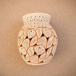 May include: A beige ceramic vase with a fish and leaf design. The vase is mounted on a textured wall. The vase has a wide body and a narrow neck with a pattern of circles. The fish and leaf design is carved into the vase.
