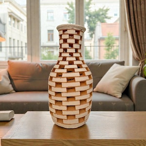 Op de afbeelding: Een decoratieve, beige en witte vaas met een uniek baksteenachtig ontwerp. De vaas is gemaakt van een lichtgekleurd materiaal en staat op een houten tafel. De vaas is ongeveer 30 cm hoog.